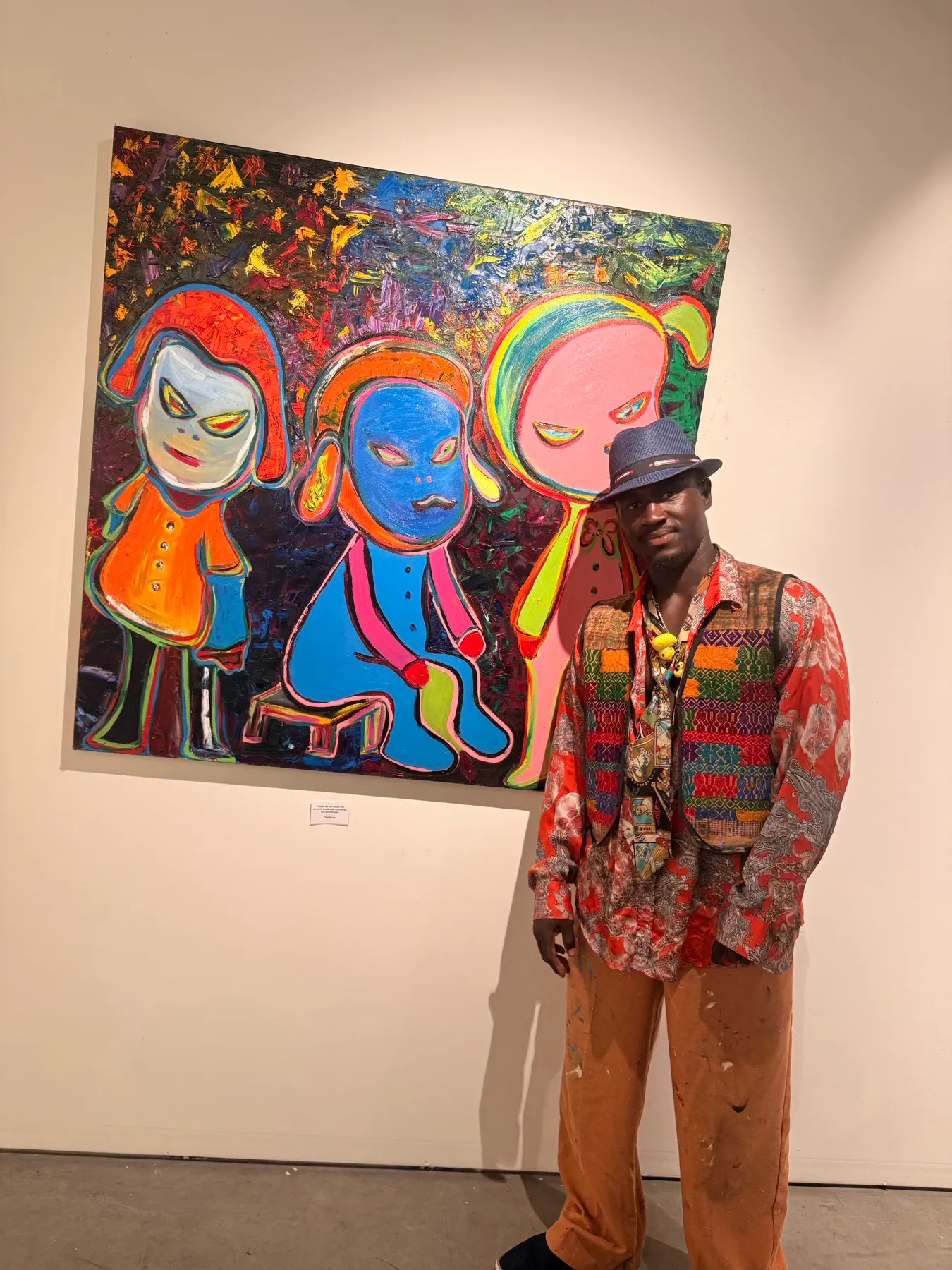 Ooowwee Walker in a fedora and woven vest standing beside his large three-figure canvas on a gallery wall with a wall label