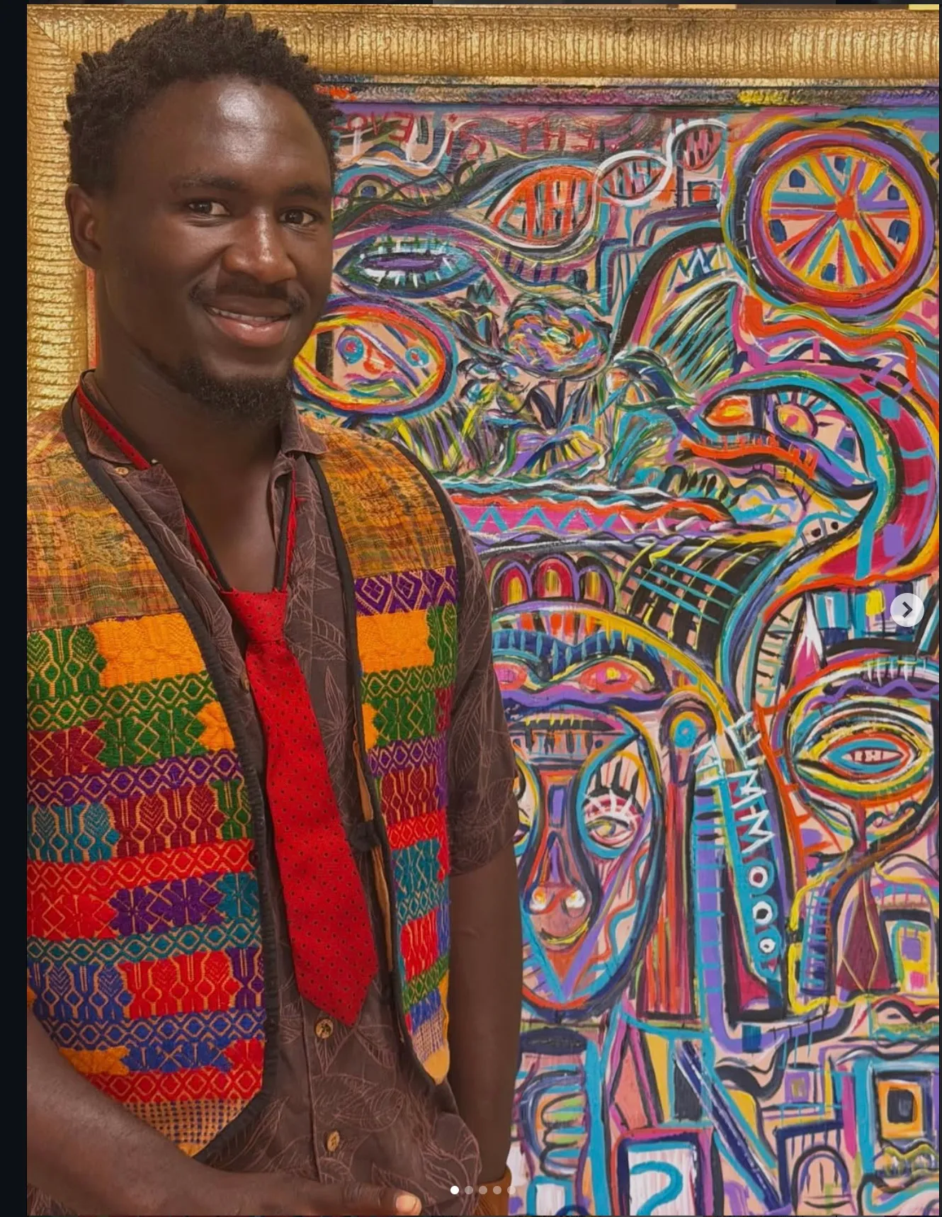Ooowwee Walker smiling in a woven vest and red tie beside one of his framed paintings