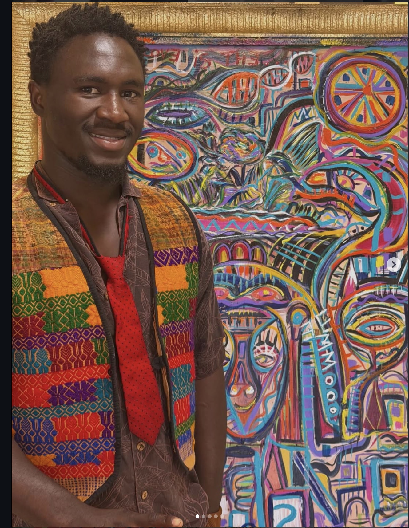 Ooowwee Walker smiling in a woven vest and red tie beside one of his framed paintings